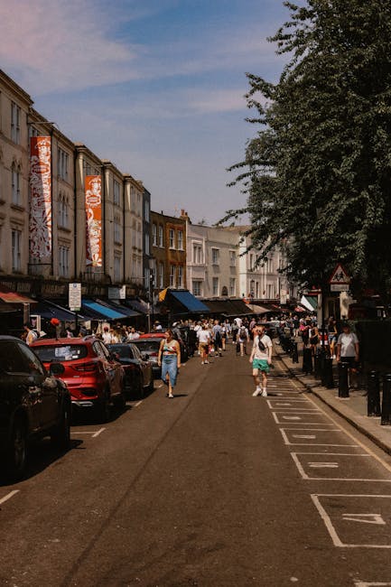 A busy street scene in Brixton with pedestrians walking along the pavement and vehicles parked on both sides of the road. In the foreground, several large cardboard boxes and pieces of packing materia