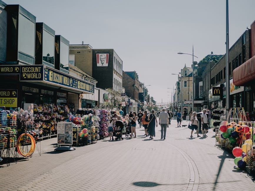 A busy street scene in Brixton with pedestrians walking along the pavement and vehicles parked on both sides of the road. In the foreground, several large cardboard boxes and pieces of packing materia