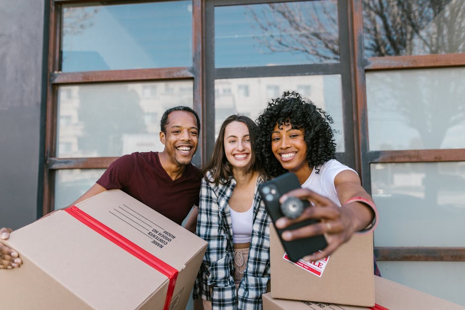 A woman with curly hair, wearing a white t-shirt and jeans, is standing at the entrance to a brick building, carrying two cardboard boxes, one in each hand, during a home relocation process. She is on